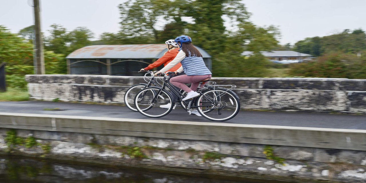 two women cycling along the grey river canal with green trees in the background